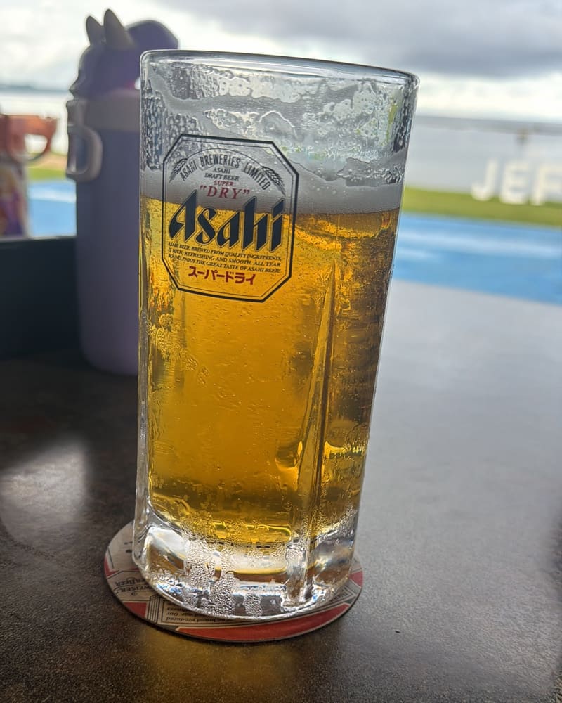 A frosty mug of Asahi Super Dry on a table at the Cove, with the Pacific and the giant JEFF'S letters behind it
