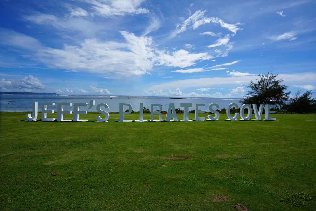 Giant white JEFF'S PIRATES COVE letters on the green lawn above Togcha Beach, with the Pacific and a blue sky behind