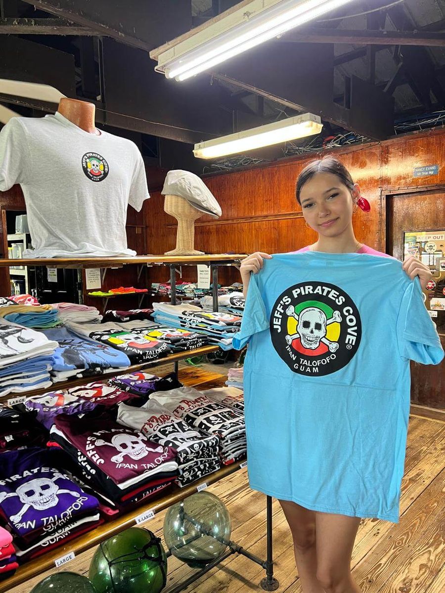 A smiling girl holds up a light-blue JPC logo tee in the wood-paneled gift shop, with stacks of skull tees and glass floats around her