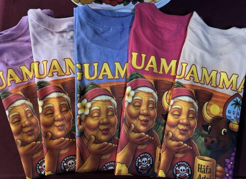 A fanned stack of Guamma tees in five colors, each printed with a smiling island grandma