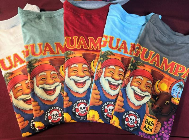 A fanned stack of Guampa tees in five colors, each printed with a smiling island grandpa