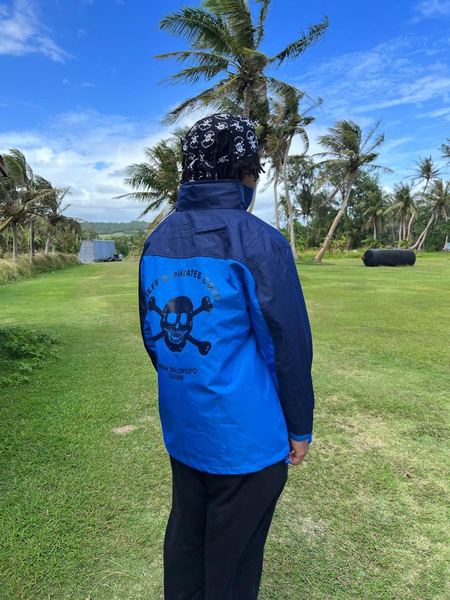 The back of a blue JPC windbreaker with the skull logo, worn on the lawn under the palms