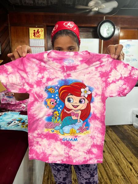 A pink tie-dye kids tee with a mermaid and the word Guam, held up in the shop