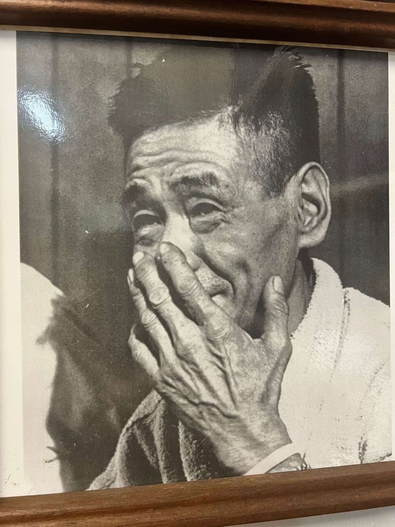 Shoichi Yokoi with a hand raised to his face, a framed black-and-white portrait
