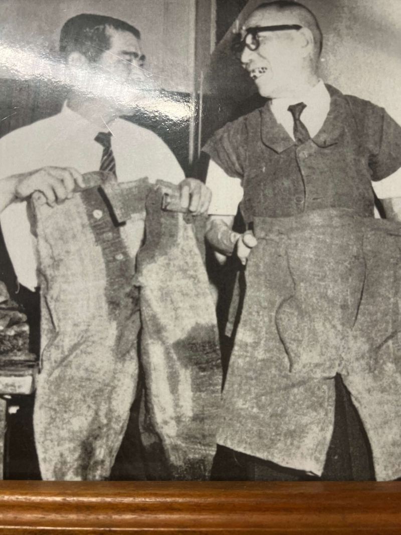 Two men in suits holding up handmade trousers and a jacket